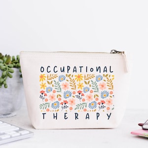 Peut inclure: Une trousse à fermeture éclair crème avec les mots "OCCUPATIONAL THERAPY" en bleu marine. La trousse présente un motif floral avec des fleurs jaunes, bleues et roses. Une plante succulente et un clavier sont en arrière-plan.