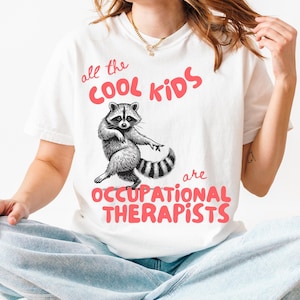 May include: White t-shirt with a raccoon graphic and the text "all the COOL KIDS are OCCUPATIONAL THERAPISTS" in red. The raccoon is black and white with a striped tail. The shirt is a casual style.