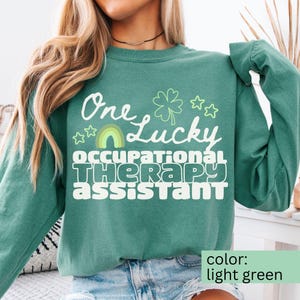 May include: Light green sweatshirt with the text "One Lucky Occupational Therapy Assistant" in white. The design includes a shamrock, stars, and a rainbow. The sweatshirt is a solid light green color.