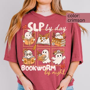 Peut inclure: T-shirt couleur cramoisi avec un motif graphique de fantômes de dessins animés lisant des livres, avec le texte "SLP by day, Bookworm by night". Le motif comprend des feuilles d'automne et des fleurs.