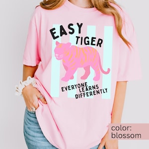 K&ouml;nnte beinhalten: Ein hellrosa T-Shirt mit einer Grafik eines rosa Tigers mit gelben Streifen. Der Text "EASY TIGER" steht &uuml;ber dem Tiger und "EVERYONE LEARNS DIFFERENTLY" ist darunter gedruckt. Das Design ist vor vertikalen hellblauen Streifen platziert.