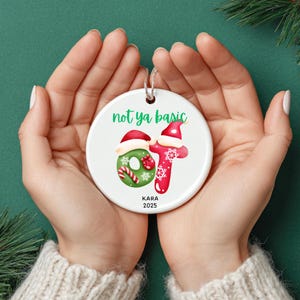 Könnte beinhalten: Ein weißer Keramik-Weihnachtsschmuck mit dem Text "not ya basic" in Grün. Der Schmuck zeigt ein festliches Design mit einem roten Weihnachtsmannhut, einem grünen Kranz und den Buchstaben "OT" in Rot mit Schneeflocken. Der Name "KARA" und das Jahr "2025" sind darunter aufgedruckt.