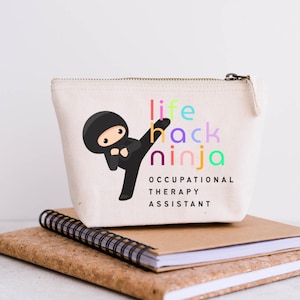 May include: A canvas zipper pouch with the words "life hack ninja" in colorful letters and a cartoon ninja kicking. Below the text are the words "Occupational Therapy Assistant". The pouch is sitting on a notebook.