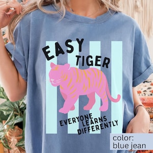 K&ouml;nnte beinhalten: Ein blaues T-Shirt in Jeansoptik mit einer Grafik eines rosa Tigers mit gelben Streifen. Der Text "EASY TIGER" steht in Schwarz &uuml;ber dem Tiger, und "EVERYONE LEARNS DIFFERENTLY" ist in Schwarz darunter auf hellblauen vertikalen Streifen gedruckt.
