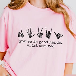 May include: Pale pink t-shirt featuring a black graphic of five skeleton hand gestures. The text "you're in good hands, wrist assured" is printed below the hands.