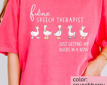 May include: A bright pink t-shirt with the text "Future Speech Therapist" in a cursive font. Below the text are five cartoon ducks, each with a unique accessory. The phrase "Just Getting My Ducks In A Row" is printed below the ducks. The shirt's color is crunchberry.