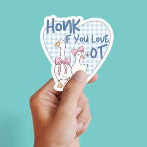 May include: A heart-shaped sticker with a blue gingham pattern. The sticker shows two cartoon geese wearing pink bows, with the text "Honk If You Love OT" in blue. The sticker is held by a hand against a teal background.