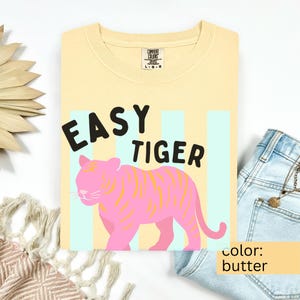 K&ouml;nnte beinhalten: Ein buttergelbes T-Shirt mit Rundhalsausschnitt und dem schwarzen Schriftzug EASY TIGER &uuml;ber einer Illustration eines rosa Tigers mit gelben Streifen. Das Design ist vor hellblauen vertikalen Streifen platziert. Das Shirt hat ein Comfort Colors Etikett.