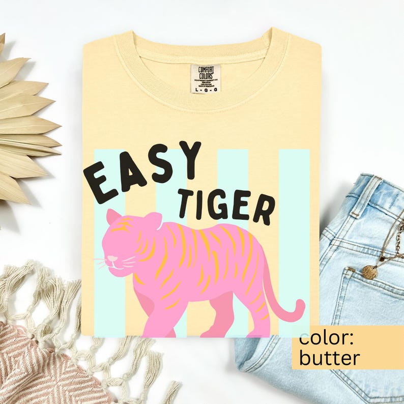 K&ouml;nnte beinhalten: Ein buttergelbes T-Shirt mit Rundhalsausschnitt und dem schwarzen Schriftzug EASY TIGER &uuml;ber einer Illustration eines rosa Tigers mit gelben Streifen. Das Design ist vor hellblauen vertikalen Streifen platziert. Das Shirt hat ein Comfort Colors Etikett.