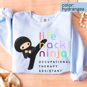 Może przedstawiać: Jasnoniebieska bluza z napisem "life hack ninja" w kolorowych literach i grafiką z kreskówkowym ninja. Tekst "OCCUPATIONAL THERAPY ASSISTANT" jest wydrukowany pod grafiką ninja. Bluza jest w kolorze hortensji.