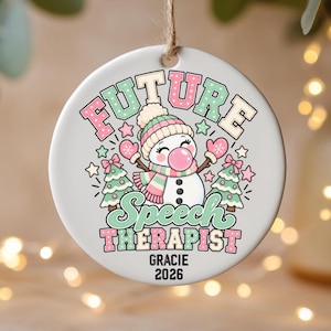 May include: A round white ceramic ornament featuring a cartoon snowman wearing a pink and green striped hat and scarf, blowing a pink bubble. The text reads FUTURE SPEECH THERAPIST, GRACIE, and 2026, surrounded by pink and green Christmas trees.