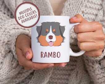 May include: White ceramic mug with a cartoon Bernese Mountain Dog illustration and the name "RAMBO" printed on the front. The mug is held by a person's hands. The text "MULTIPLE DOG COLORS AVAILABLE!" is in a circle above the mug.