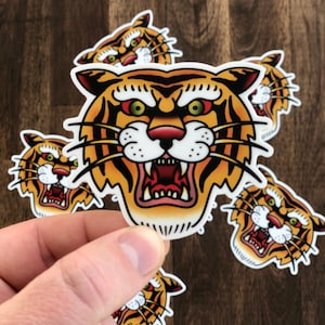 May include: Five colorful tiger head stickers with a traditional tattoo style. The tiger has orange and black stripes, a white face, and red eyes.