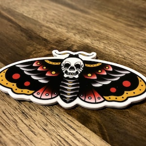 Puede incluir: Una polilla negra y blanca con acentos rojos y amarillos y una calavera en el centro. La polilla es un dise&ntilde;o de tatuaje tradicional.