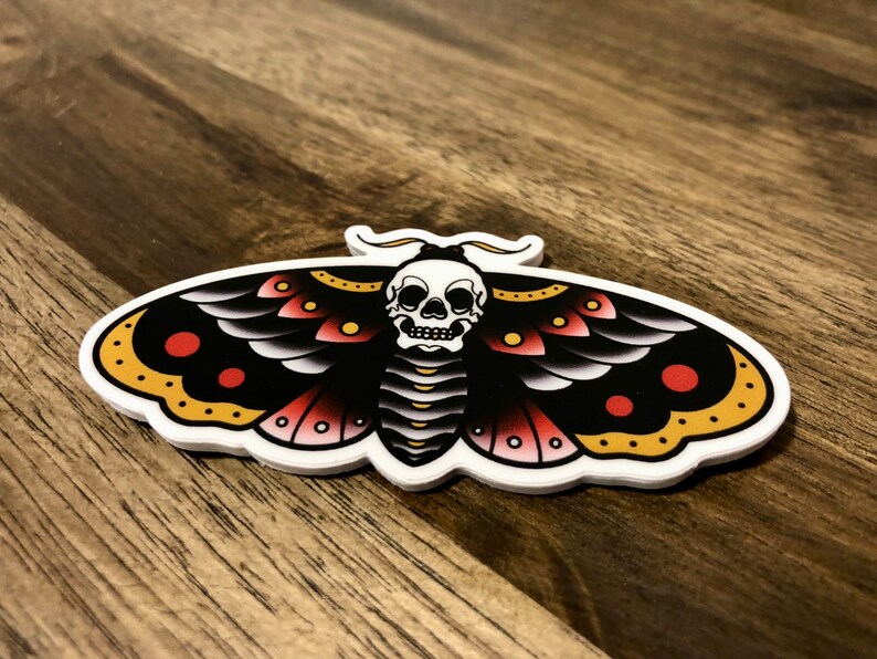 Puede incluir: Una polilla negra y blanca con acentos rojos y amarillos y una calavera en el centro. La polilla es un dise&ntilde;o de tatuaje tradicional.