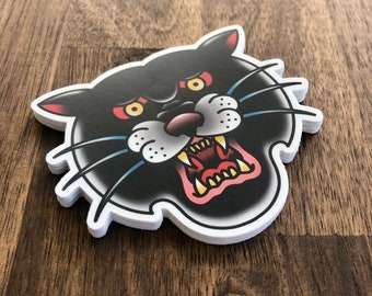 May include: A black and white sticker of a snarling panther with yellow eyes. The sticker has a white border and is printed on a glossy surface.