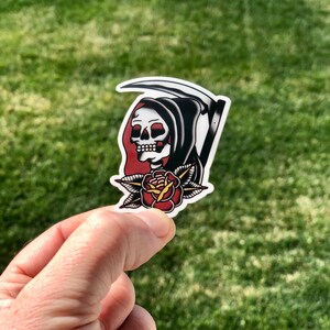 May include: A black and white sticker of the Grim Reaper with a red rose. The Grim Reaper is wearing a black hooded robe and holding a scythe. The rose is red with green leaves.