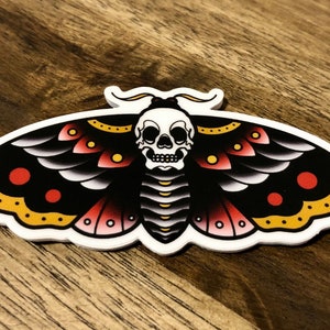 Puede incluir: Una polilla negra y blanca con acentos rojos y amarillos. La polilla tiene una calavera por cabeza y es un dise&ntilde;o de tatuaje tradicional.