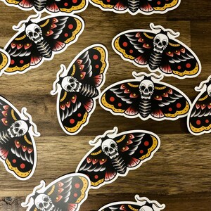 Puede incluir: Un conjunto de diez pegatinas de polilla negras y blancas con acentos rojos y amarillos. Cada polilla tiene un dise&ntilde;o de calavera en su cuerpo.