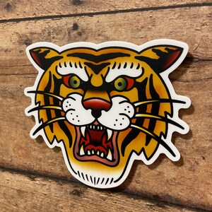 May include: A traditional tattoo style illustration of a tiger's face with a fierce expression. The tiger has orange and black stripes, a white muzzle, and green eyes.