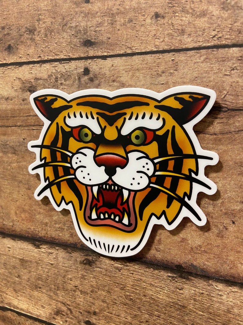 May include: A traditional tattoo style illustration of a tiger's face with a fierce expression. The tiger has orange and black stripes, a white muzzle, and green eyes.