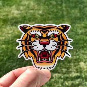 May include: A close-up of a tiger face sticker. The sticker is yellow, orange, and black with green eyes and a red nose. The tiger is snarling with its mouth open.
