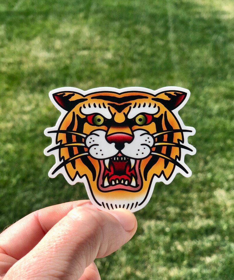 May include: A close-up of a tiger face sticker. The sticker is yellow, orange, and black with green eyes and a red nose. The tiger is snarling with its mouth open.