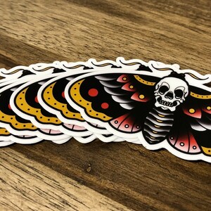 Puede incluir: Un conjunto de ocho pegatinas de polilla negra y amarilla con un dise&ntilde;o de calavera. La polilla tiene detalles rojos y blancos en sus alas.
