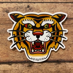 May include: A traditional tattoo style illustration of a tiger's face. The tiger has orange and black stripes, a white muzzle, and a fierce expression with bared teeth. The background is a wood grain texture.