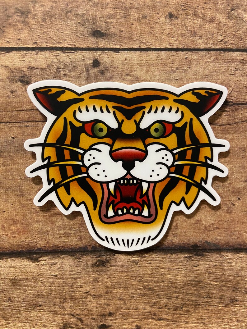 May include: A traditional tattoo style illustration of a tiger's face. The tiger has orange and black stripes, a white muzzle, and a fierce expression with bared teeth. The background is a wood grain texture.