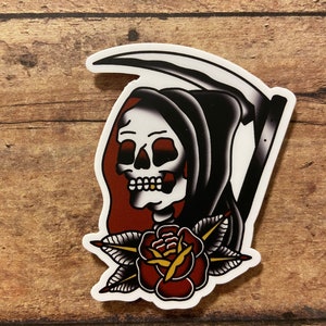 May include: A traditional tattoo style sticker featuring a black and white skeletal figure with a red hooded robe, holding a black scythe. The figure is holding a red rose with green leaves and a gold center.