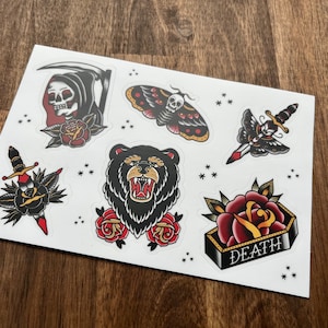 May include: A sheet of traditional tattoo flash stickers featuring a black bear, a skull with a scythe, a dagger with a rose, a moth, a dagger with a butterfly, and a rose with the word "DEATH" in a banner.