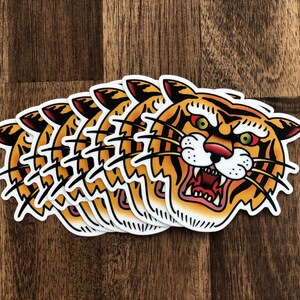 May include: A set of five tiger head stickers with a traditional tattoo style. The tiger has orange and black stripes, a white face, and a red mouth with sharp teeth. The stickers are printed on white paper.