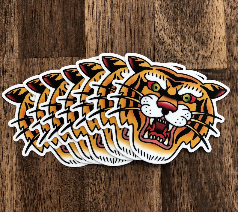 May include: A set of five tiger head stickers with a traditional tattoo style. The tiger has orange and black stripes, a white face, and a red mouth with sharp teeth. The stickers are printed on white paper.