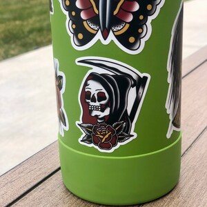 May include: Green reusable water bottle with various colorful stickers, including a butterfly, a grim reaper with a scythe, and a rose.