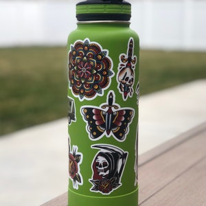 May include: A green reusable water bottle with various colorful stickers, including a mandala, a butterfly, a skull with a dagger, and a grim reaper.