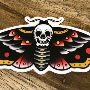 Puede incluir: Una polilla negra y roja con una calavera blanca en el centro. La polilla tiene acentos amarillos y puntos rojos en sus alas.