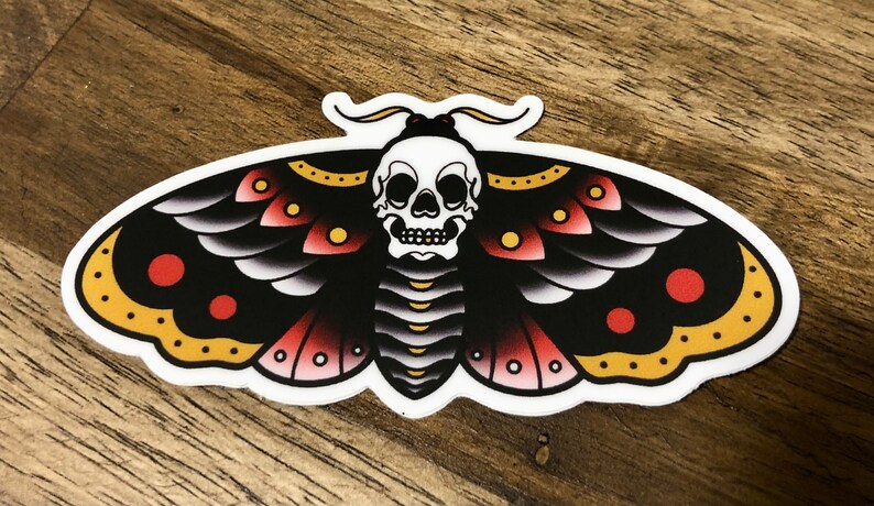 Puede incluir: Una polilla negra y roja con una calavera blanca en el centro. La polilla tiene acentos amarillos y puntos rojos en sus alas.