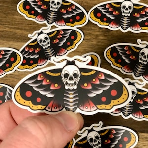 Puede incluir: Un conjunto de ocho pegatinas de polillas negras y rojas con una calavera blanca y detalles amarillos. Las polillas tienen un estilo de tatuaje tradicional.
