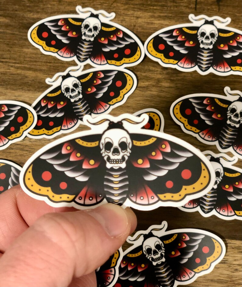 Puede incluir: Un conjunto de ocho pegatinas de polillas negras y rojas con una calavera blanca y detalles amarillos. Las polillas tienen un estilo de tatuaje tradicional.