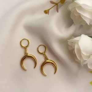 May include: A pair of gold-colored crescent moon earrings. Each earring features a small hoop that connects to a chain, which then connects to the crescent moon. The earrings are displayed on a white, silky surface.