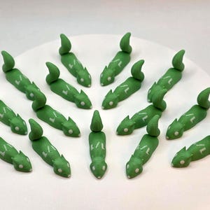 May include: A collection of green, lizard-shaped figurines with white accents, arranged on a white surface. Each figurine has a curved tail and a detailed body. The figurines are likely handmade and could be used as decorative items or cake toppers.