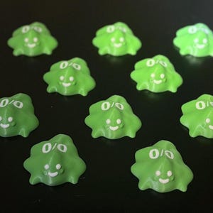 May include: A collection of ten small, lime green, blob-shaped objects with white smiling faces and eye details. The objects are scattered on a dark surface, creating a playful and whimsical arrangement.