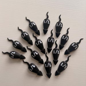May include: A collection of black mouse-shaped objects with curved tails and the text "L/L" in white. The objects are arranged on a light-colored surface. The objects appear to be decorative or possibly game pieces.