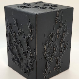 May include: A black, cube-shaped box with a textured, raised design on its surfaces. The design appears to be an abstract, organic pattern. The box is sitting on a white surface.
