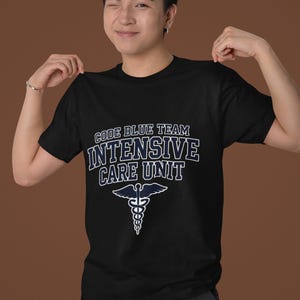 Puede incluir: Una camiseta negra de manga corta con texto blanco que dice CODE BLUE TEAM INTENSIVE CARE UNIT sobre un símbolo médico de caduceo blanco. El diseño está centrado en el pecho de la prenda, que tiene un cuello redondo y un ajuste estándar.