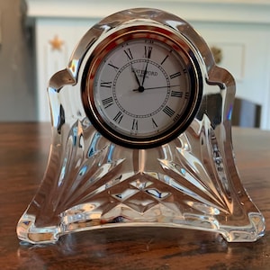 Puede incluir: Un reloj Waterford de cristal transparente con una esfera redonda con números romanos y el nombre de la marca. El reloj tiene un bisel dorado y una base decorativa facetada. El reloj está sobre una superficie marrón.