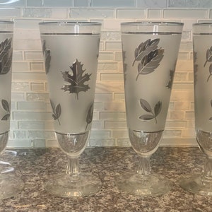 May include: Set of four frosted glass goblets with silver leaf designs. The goblets have a clear glass base and a silver rim.