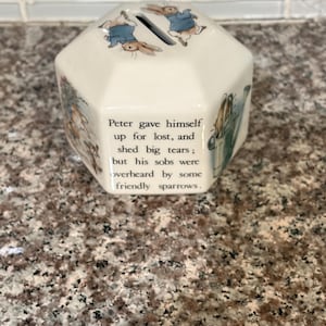 May include: Cream-colored, multi-sided ceramic piggy bank featuring illustrations of rabbits in blue jackets. The bank has a slot on top and text that reads, "Peter gave himself up for lost, and shed big tears; but his sobs were overheard by some friendly sparrows."