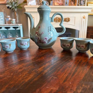 May include: A celadon-colored tea set with a teapot and six matching cups. The teapot has a curved handle and spout, and the cups are arranged in a row. Each piece is decorated with pink floral designs.